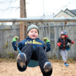 Two young children swing on swings wearing winter hats and sweatshirts