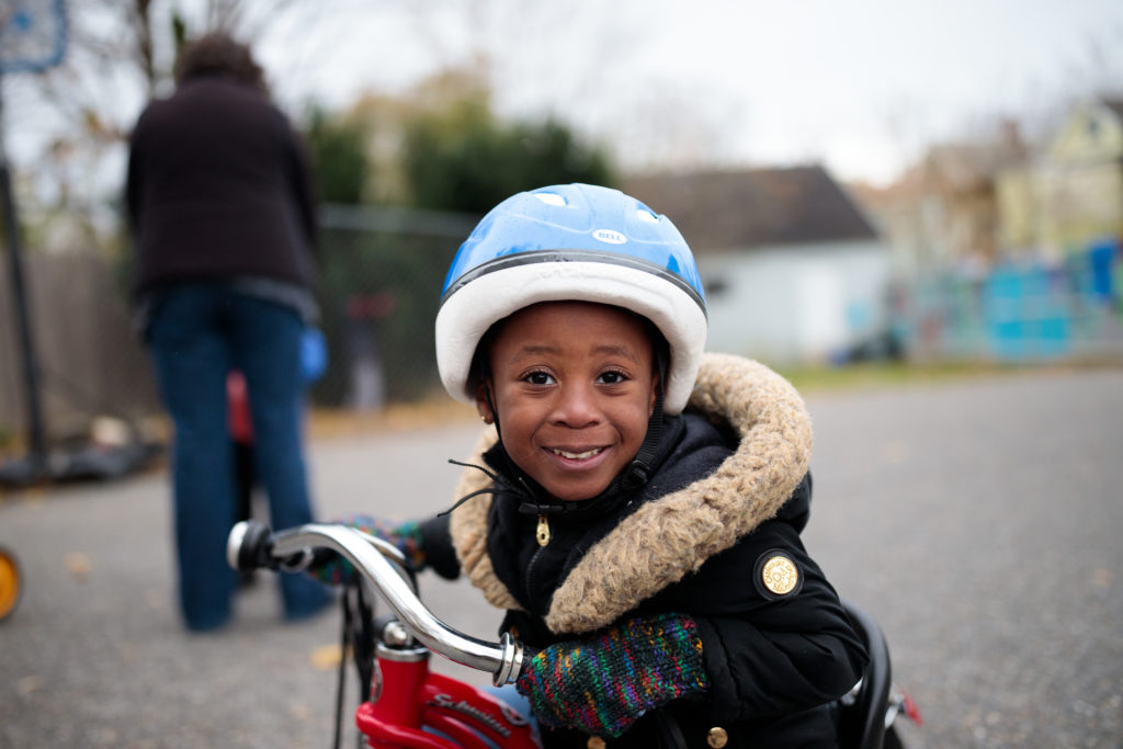 A young Black child smiles while riding a tricycle and wearing a winter jacket and helmet