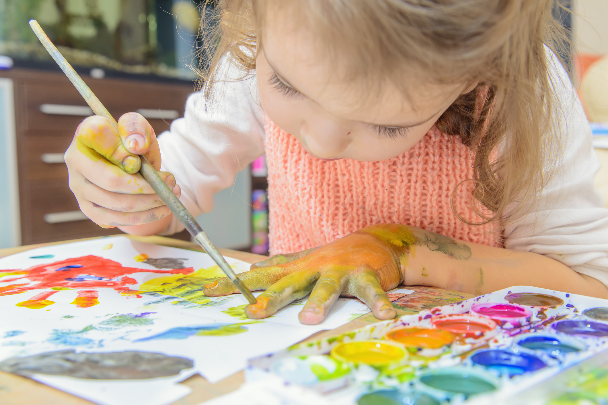 A young white girl with blond hair paints on her had and on paper