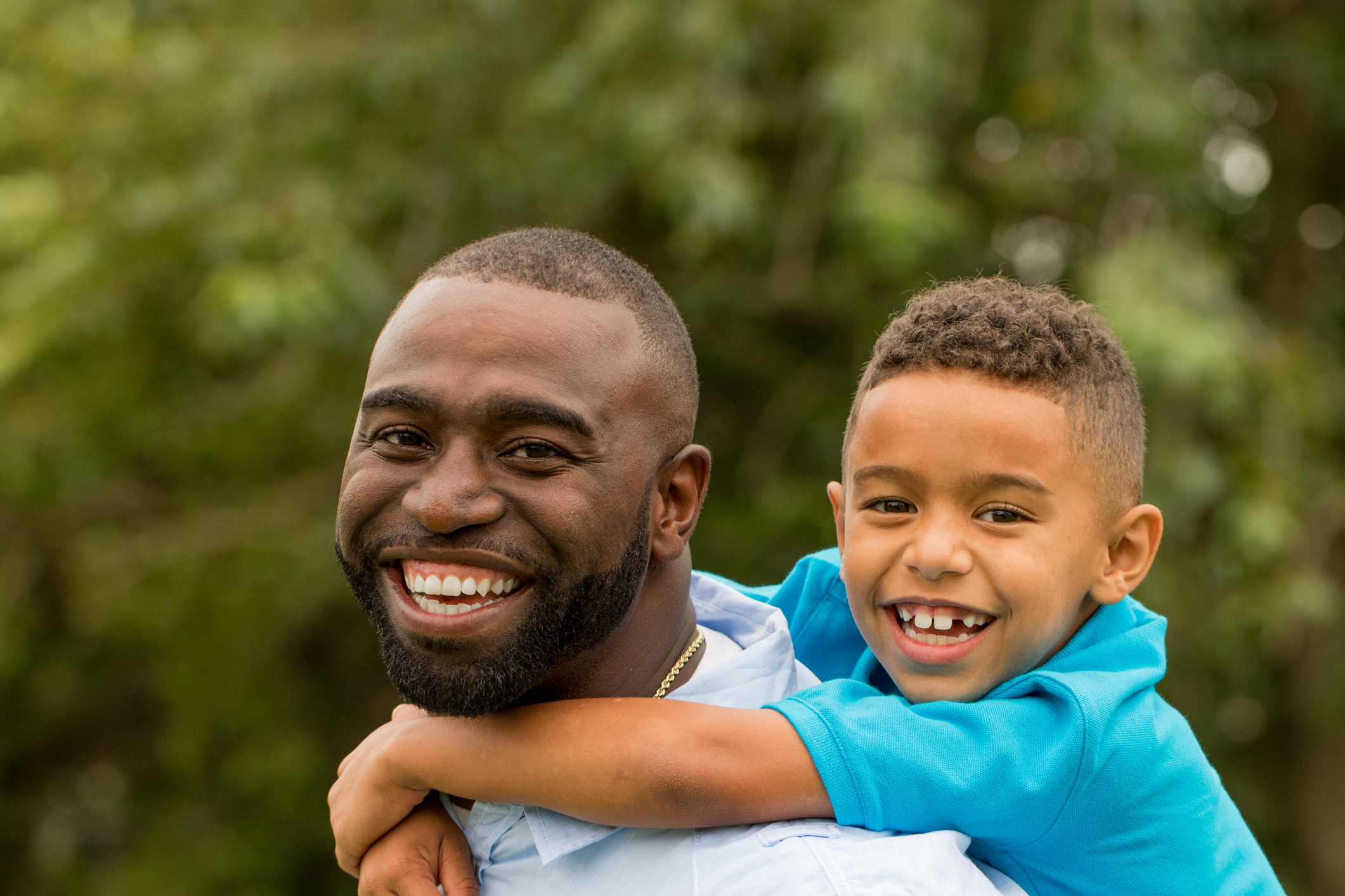 A Black dad smiles warmly while carrying his young son piggy-back