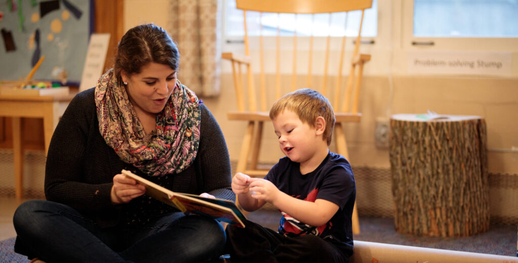 A woman reads a book to a young child while both sit on the floor