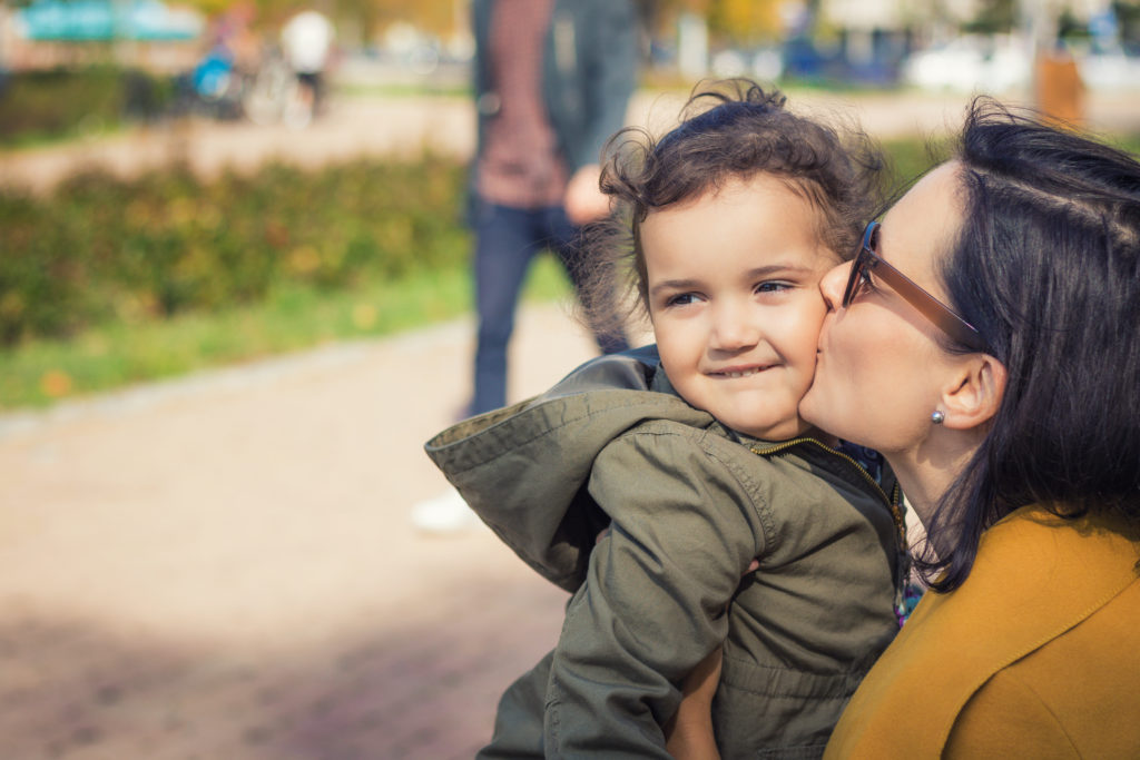 A mom with dark hair and sunglasses kisses her young daughter on the cheek