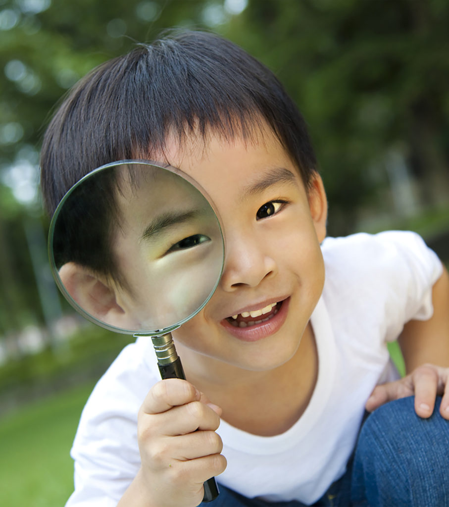 A young Asian boy smiles and looks through a magnifying glass