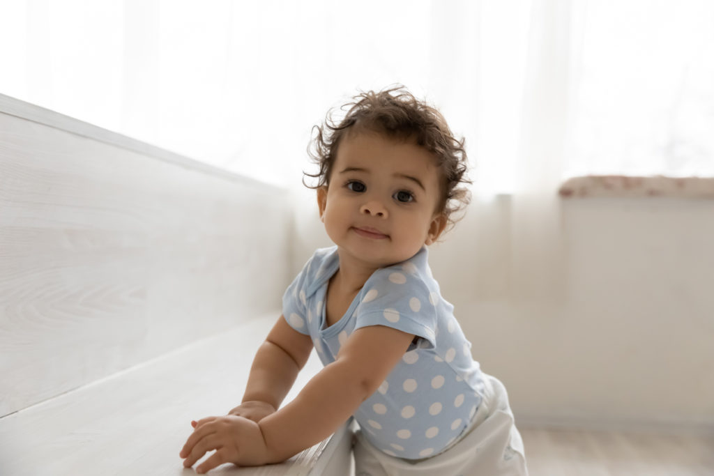 A baby with dark hair and light brown skin leans on a bench to stand up