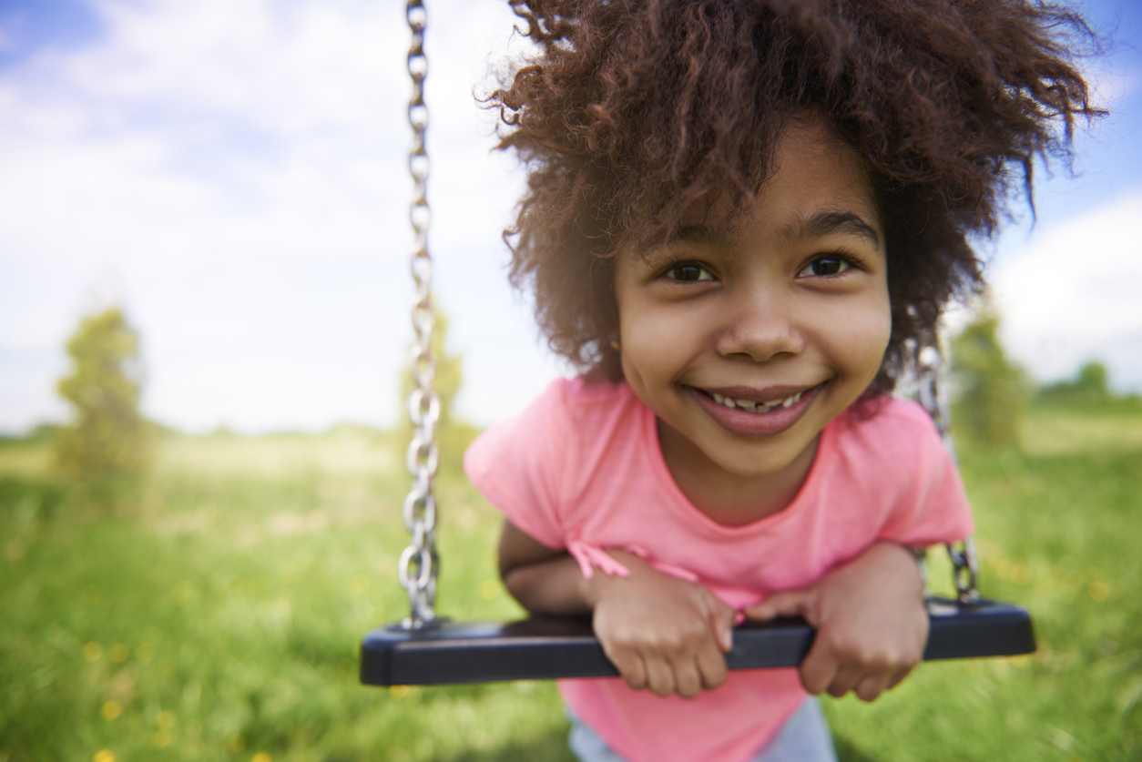 A young Black girl wearing a pink shirt leans on a swing and smiles happily