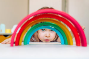 A young white girl with light brown hair looks through a rainbow made of stacked blocks