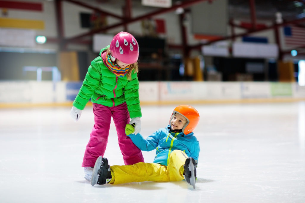 Two young children are ice skating, one offering her hand to help the other up