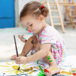 A young white girl with brown hair paints on a pad of paper