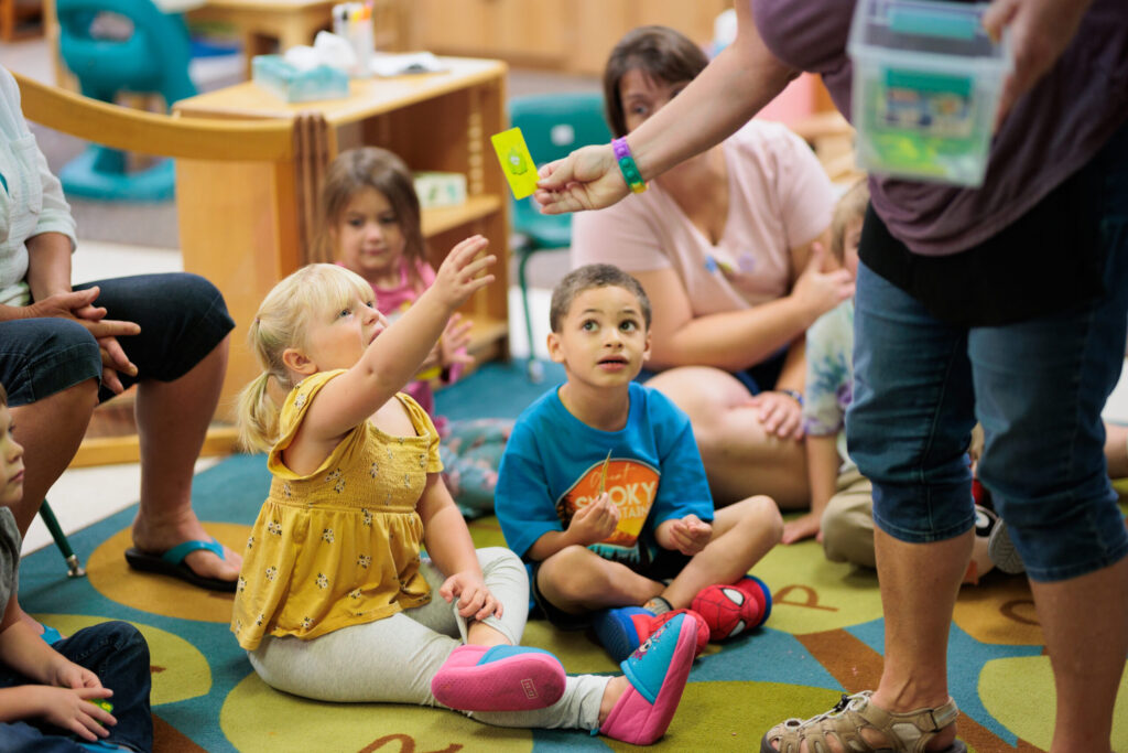 A group of young children at Gilman Head Start with one blonde child reaching to take a card from a teacher