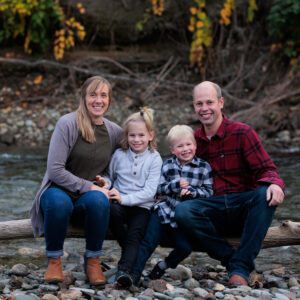 A light-skinned family with a mother and father and two young children sit together on a log, smiling