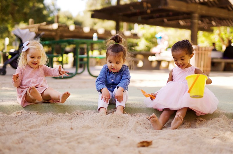 Three young children wearing colorful dresses and shirts play in a sandbox together