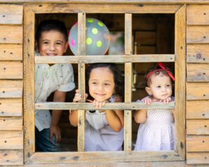 Three young children smile and look through the window of a wooden playhouse, with a polka dot balloon behind them