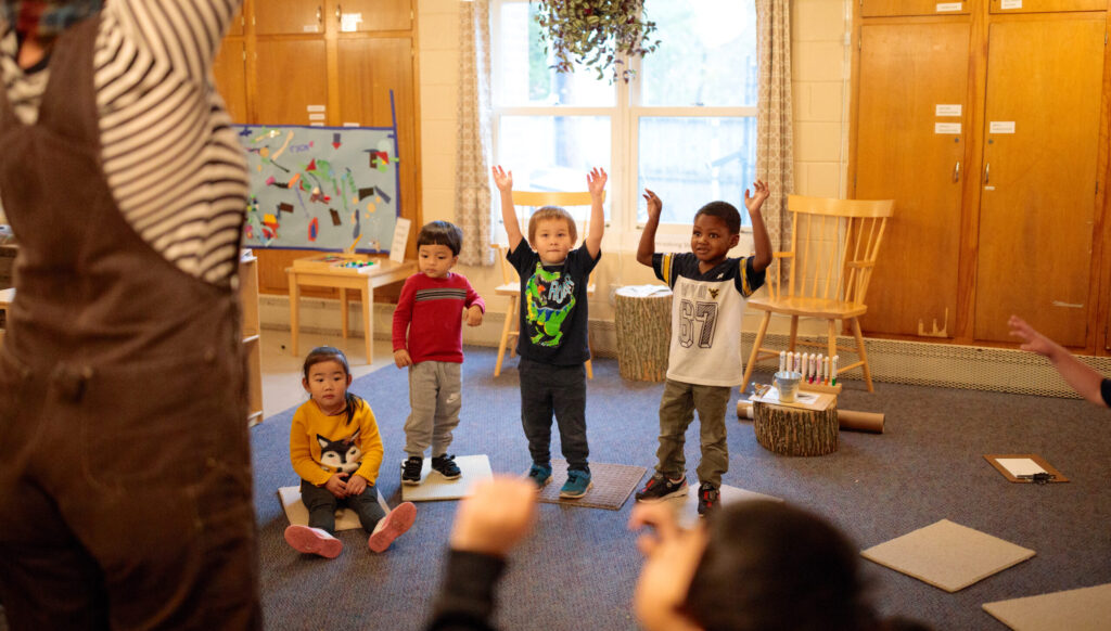 A group of young kids raise their arms while playing a game with their teacher