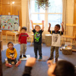 A group of young kids raise their arms while playing a game with their teacher