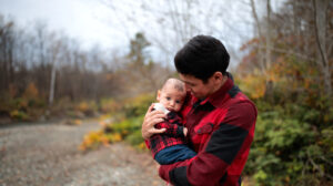 A young father with black hair holds his small baby in his arms and looks down at him lovingly