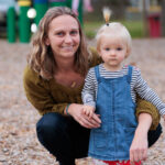 A mom smiles with her arms around her toddler who wears a denim dress and has blonde hair