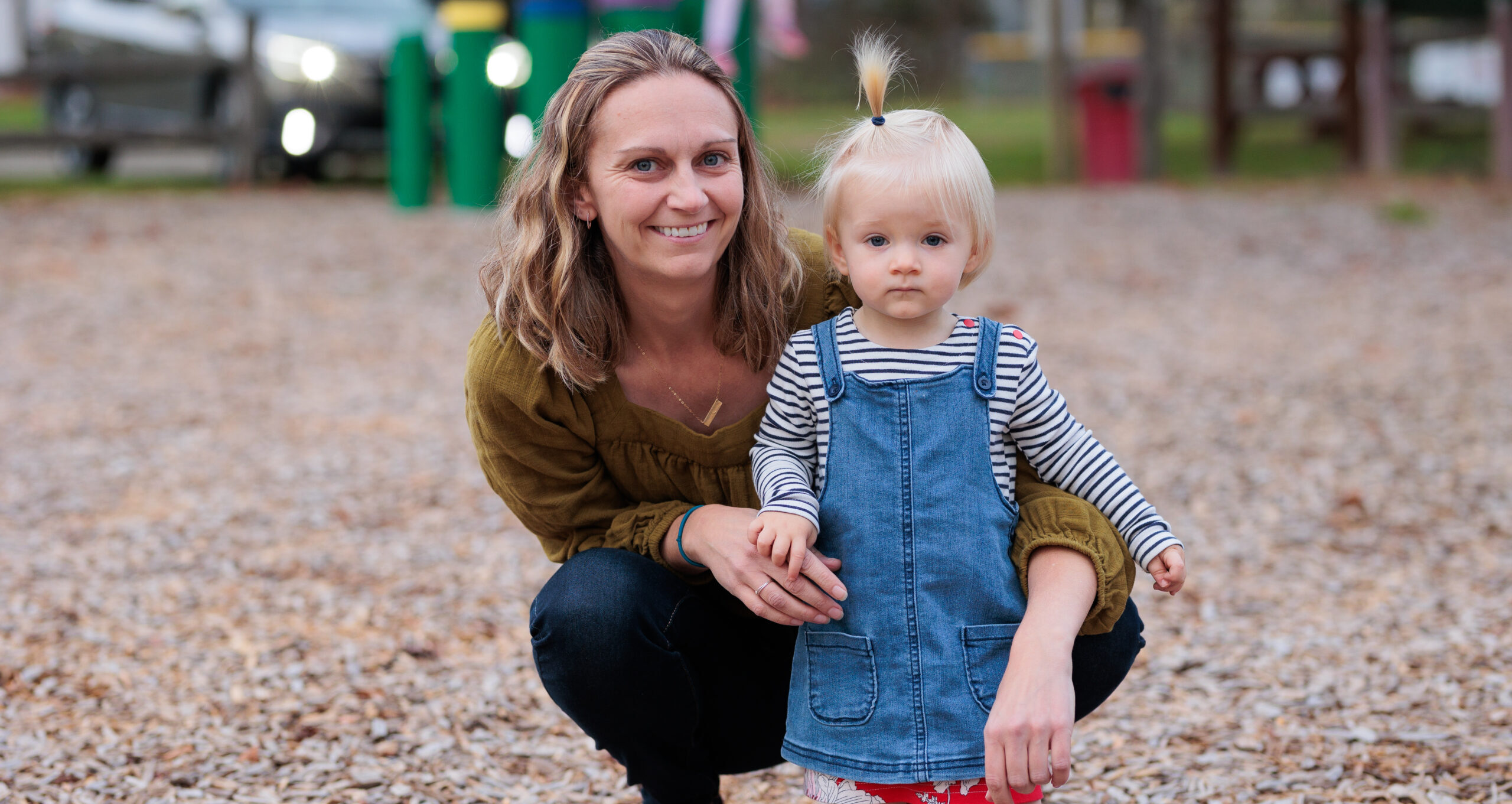 A mom smiles with her arms around her toddler who wears a denim dress and has blonde hair