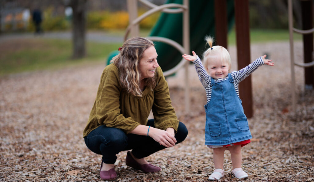 A light-skinned mom and her toddler on a playground--the mom crouches down and smiles as the toddler holds up their arms and grins