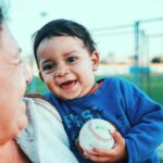 A young Latino boy smiles while holding a baseball and being carried by a caregiver