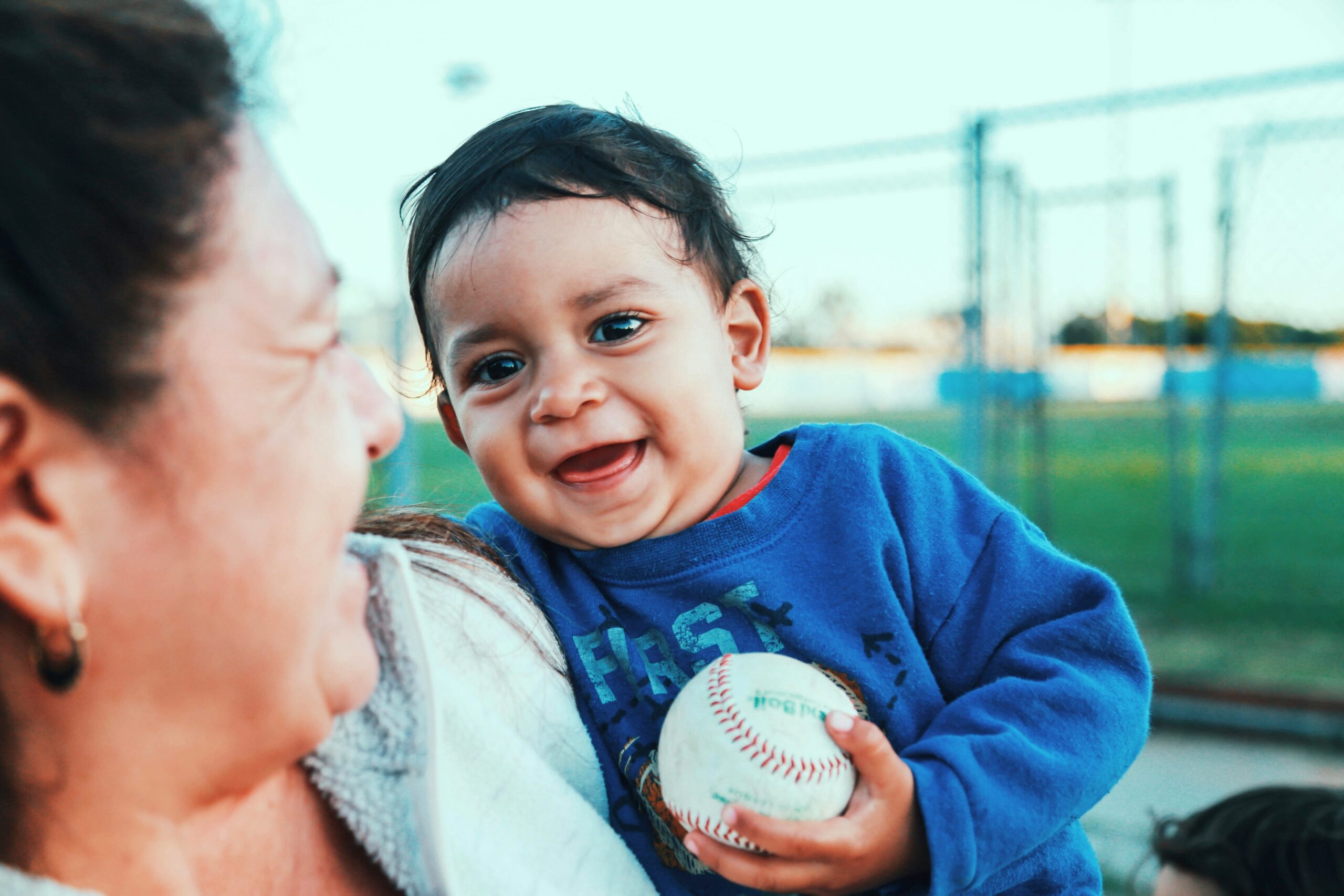 A young Latino boy smiles while holding a baseball and being carried by a caregiver