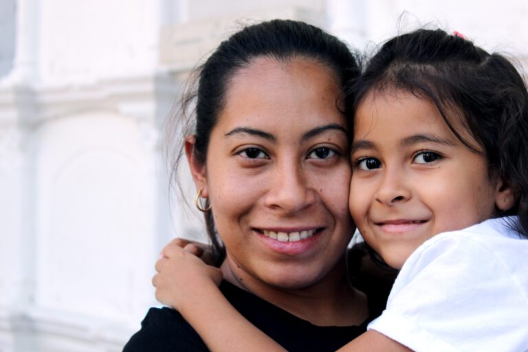 A mom with dark hair and brown skin hugs her young daughter while both smile happily