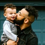 A Black dad with glasses holds his happy toddler in his arms