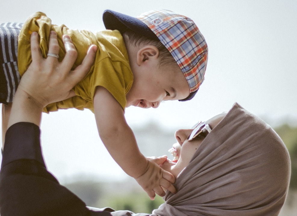 A mom wearing a head scarf and sunglasses joyfully holds her baby up over her head