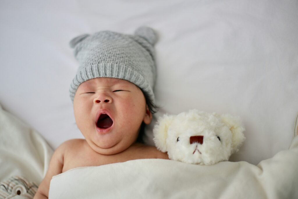 A very young baby yawns while wearing a gray hat and tucked in next to a white teddy bear