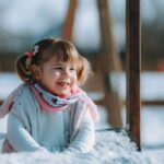 A young white girl with brown pigtails smiles while sitting in the snow