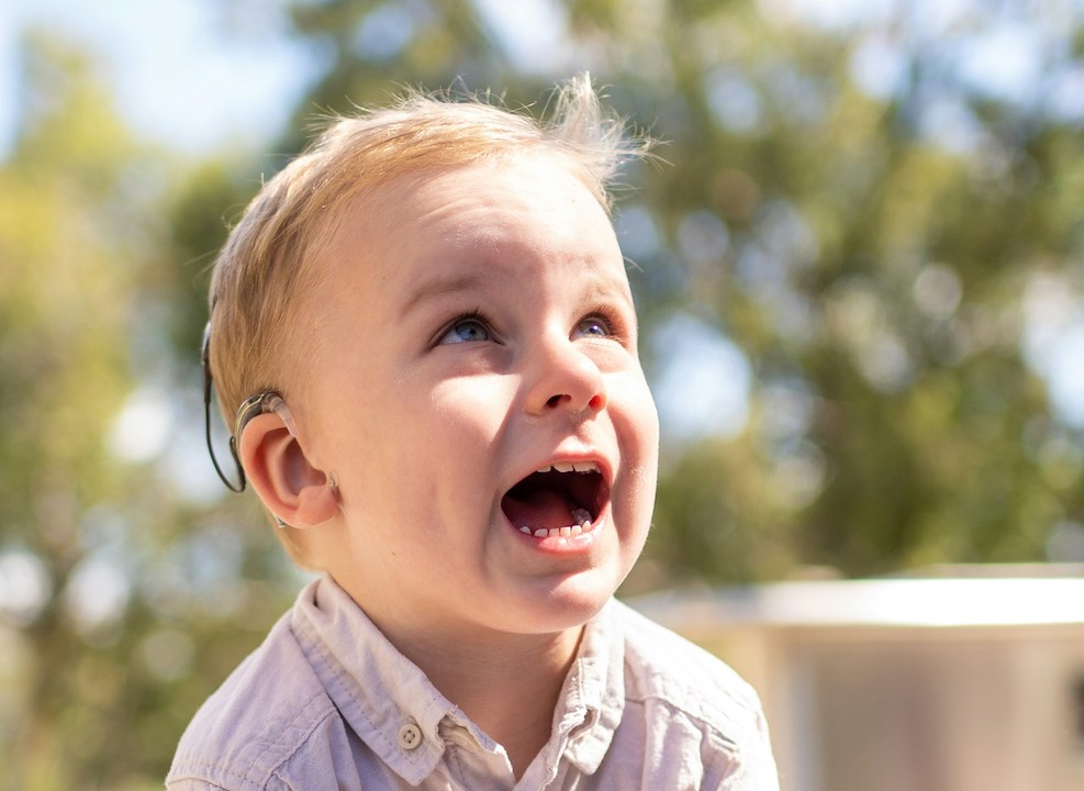 A white toddler with a hearing aid and blond hair smiles or talks joyfully