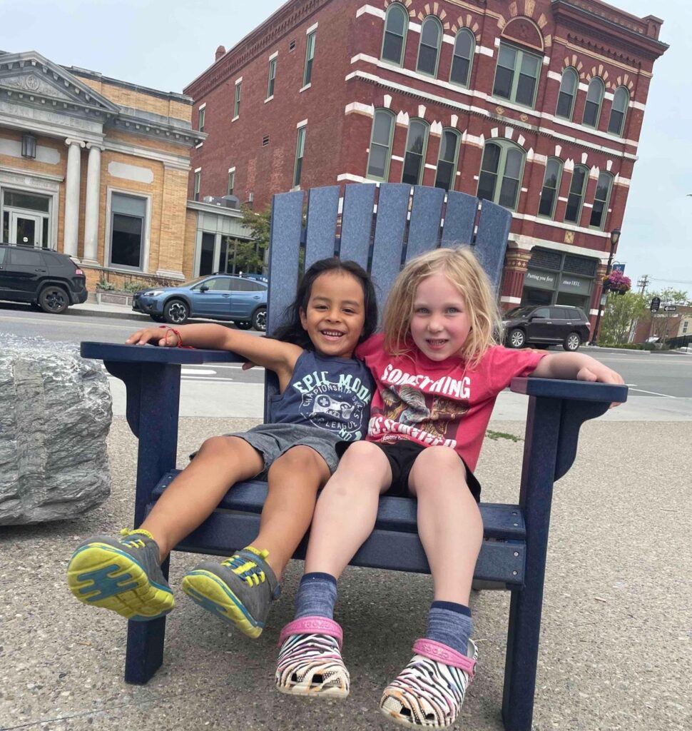 Two smiling girls sit in a chair together by the main street of Middlebury, Vermont