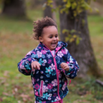 A young girl with light brown skin and dark hair runs joyfully outdoors