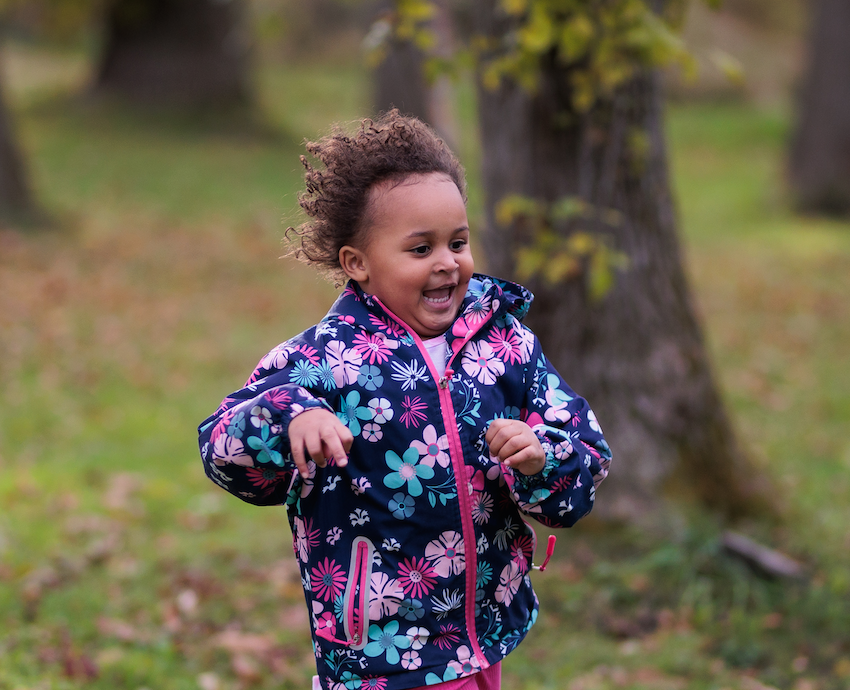 A young girl with light brown skin and dark hair runs joyfully outdoors