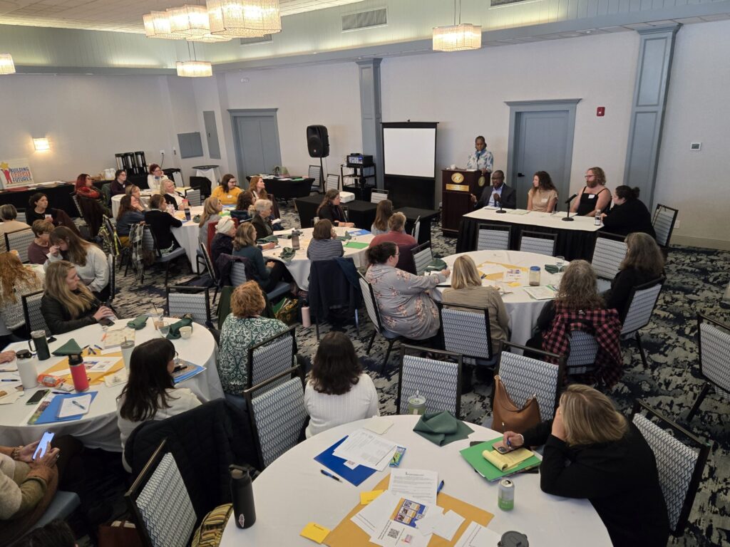 A speaker at a podium and a diverse group of four panelists at a table face a room full of Regional Council Summit attendees at round tables