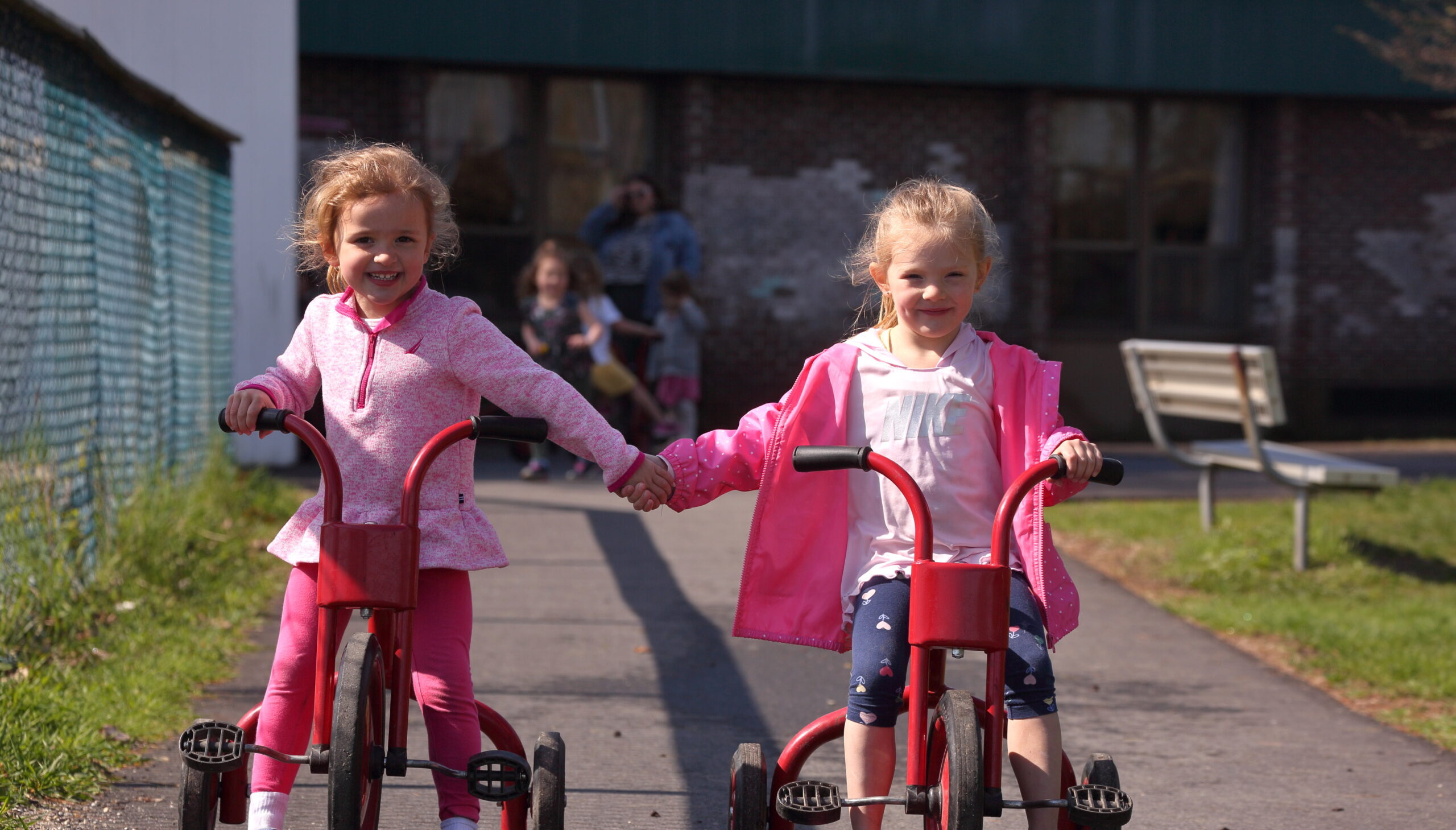 Two young girls with blonde hair and light skin ride tricycles while holding hands