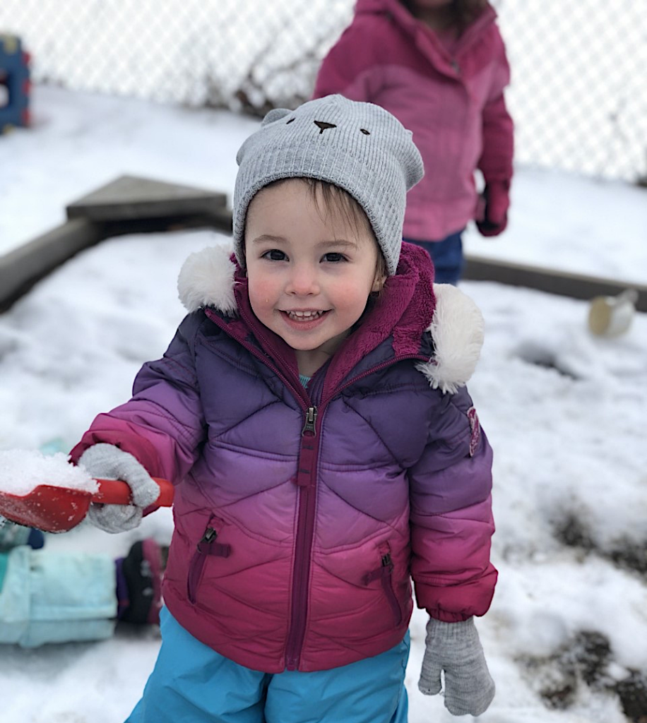 A smiling toddler in a puffy jacket plays in the snow