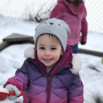 A smiling toddler in a puffy jacket plays in the snow