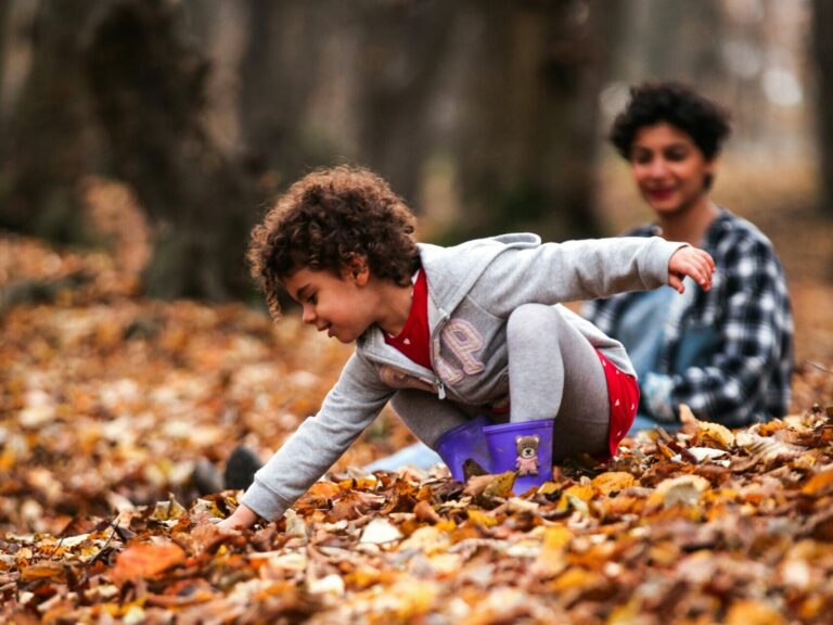 Young child smiling and reaching into dry autumn leaves as an adult looks on in the background