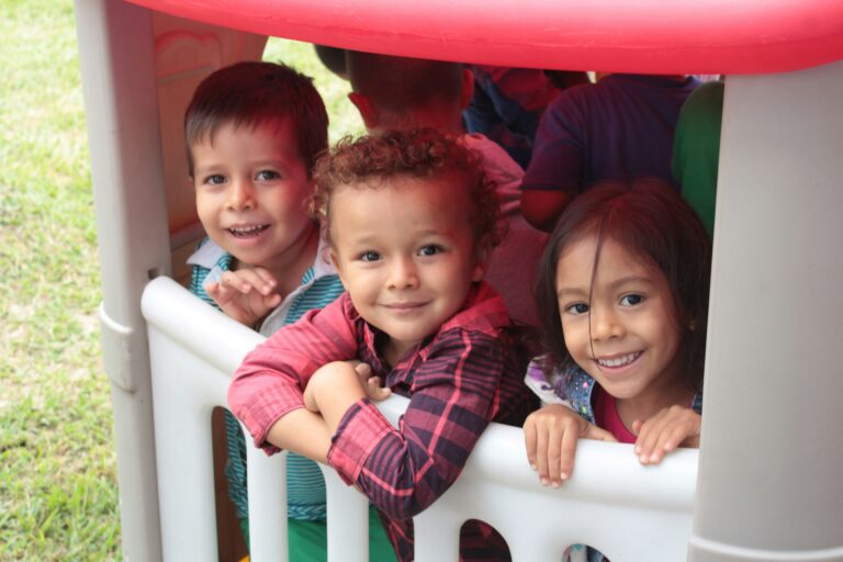 Three young children with brown hair and light brown skin smile while playing in a playhouse