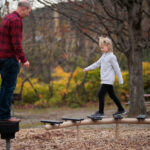 An 8 year old child with blond hair and a grey sweater looks down and balances on a beam at a playground.