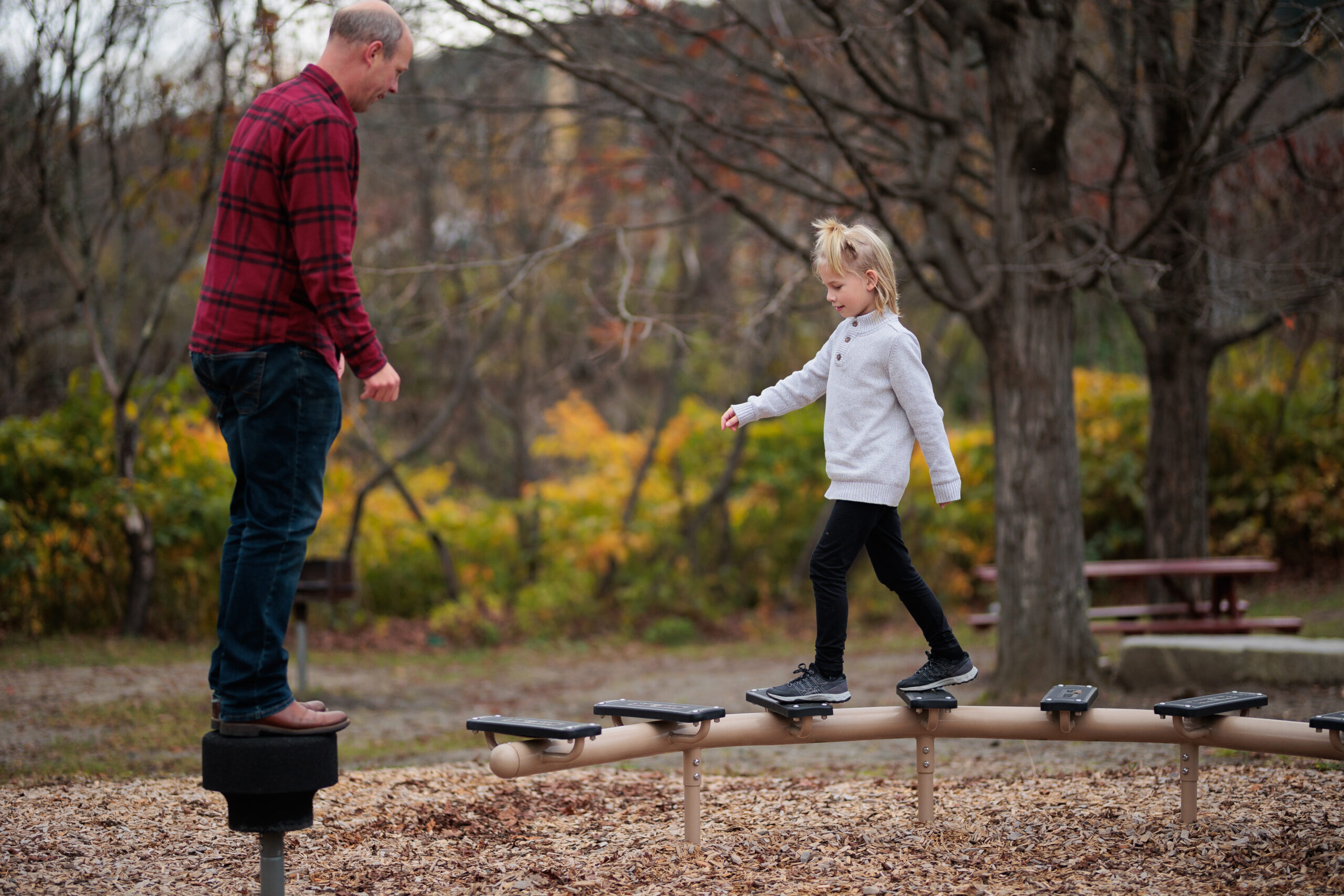 An 8 year old child with blond hair and a grey sweater looks down and balances on a beam at a playground.