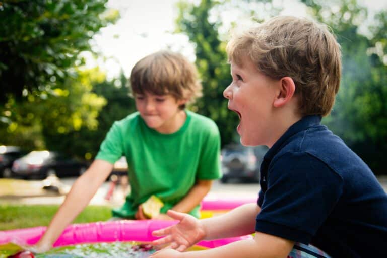 A young child with a joyful expression, and another child in the background playing with a water toy