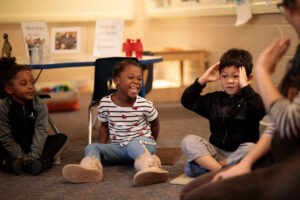 Seated preschooler, laughing preschooler, and gesturing preschooler with teacher in foreground