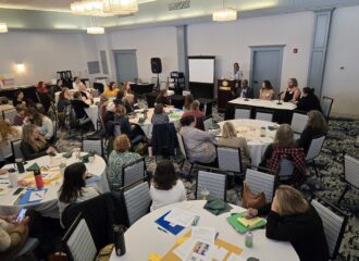 A speaker at a podium and a diverse group of four panelists at a table face a room full of Regional Council Summit attendees at round tables