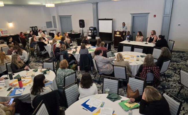 A speaker at a podium and a diverse group of four panelists at a table face a room full of Regional Council Summit attendees at round tables