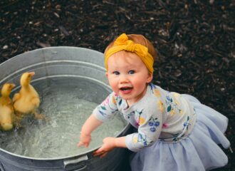 Toddler wearing a dress & yellow headband splashes in a tub where two ducklings swim