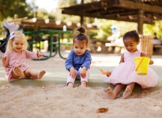 Three young children wearing colorful dresses and shirts play in a sandbox together