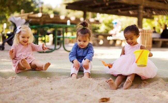 Three young children wearing colorful dresses and shirts play in a sandbox together