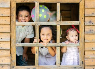 Three young children smile and look through the window of a wooden playhouse, with a polka dot balloon behind them