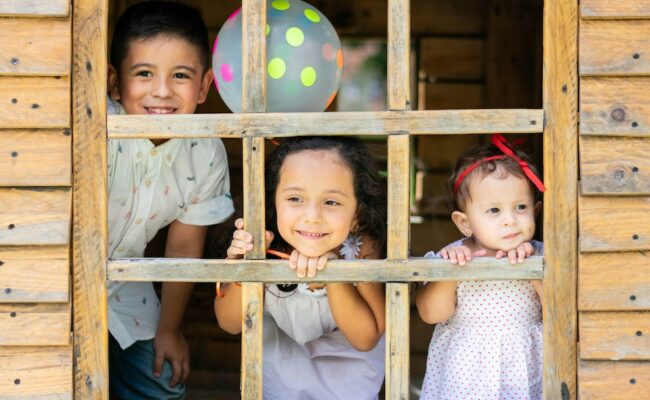 Three young children smile and look through the window of a wooden playhouse, with a polka dot balloon behind them
